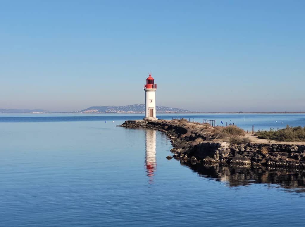Phare Méditerranéen sur Eau Calme et Arrière-plan Phare blanc à toiture rouge sur une digue rocheuse, reflété dans l'eau calme, avec une ville côtière en arrière-plan.